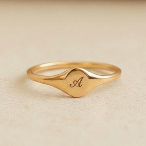 May include: A gold ring featuring an oval face with the letter "A" inscribed in a cursive script. The ring is displayed on a neutral surface. The ring's design is simple and elegant.