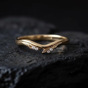 Peut inclure: Une bague dorée au design incurvé, ornée de trois petites pierres précieuses claires de chaque côté. La bague a une forme organique inspirée de la nature et est posée sur un fond sombre et texturé.