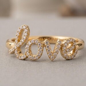 May include: A gold ring featuring the word "love" in a cursive script, embellished with small, clear gemstones. The ring's design is accentuated against a soft, neutral backdrop. The ring is a piece of jewellery.