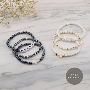 May include: Two sets of beaded bracelets. One set is dark blue with the word "MOMMY" spelled out in white letter beads, and a white cross charm. The other set is white with gold beads and the word "MOMMY" spelled out in gold letter beads, and a white cross charm.