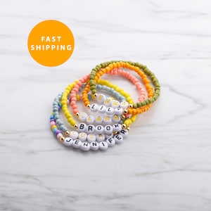 May include: A collection of colorful beaded bracelets. Each bracelet features a different color scheme, including yellow, pink, green, and blue. White letter beads spell out names. Gold-colored beads separate the names. The image includes a yellow circle with the words "FAST SHIPPING".