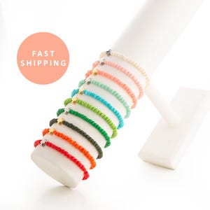 May include: A collection of colorful beaded bracelets with small cross charms. The bracelets are displayed on a white cylindrical stand. The bracelets come in various colors, including red, orange, green, blue, and pink.