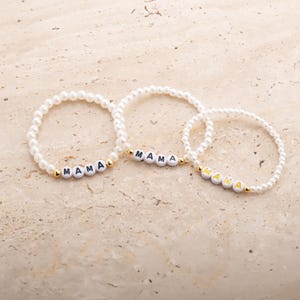 May include: Three white beaded bracelets with the word "MAMA" spelled out in white letter beads. The bracelets have gold-colored accents and are arranged on a light beige surface.