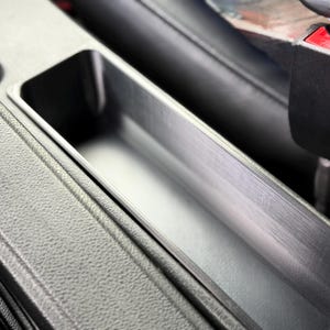 Puede incluir: Primer plano de la consola central de un coche con un compartimento rectangular. El compartimento es de color gris claro con una superficie texturizada. El entorno es gris oscuro.