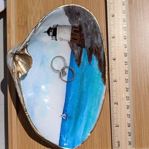 May include: Hand-painted seashell ring dish with a lighthouse scene. The dish is white with a gold rim and features a blue ocean, a lighthouse, and a small sailboat. The dish is approximately 4 inches long.