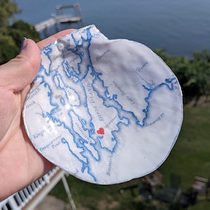 May include: A white seashell with a blue map of Rhode Island, including Newport and a red heart. The shell is held up against a blurred background of water and greenery.