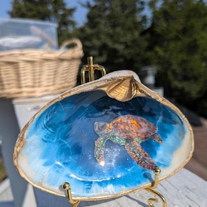 May include: A decorative seashell with a painted ocean scene and a sea turtle. The shell is edged with gold and sits on a small gold stand. The ocean is painted in shades of blue and white.