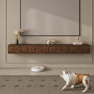 May include: A dark brown wooden floating media console with a textured, grid-patterned front. The console is mounted on a beige wall beneath a large rectangular frame. A white robotic vacuum sits on the floor near a brown and white bulldog figurine.