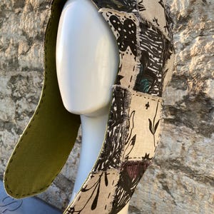 May include: A patchwork hood with a long, olive green lining. The hood is made of various fabric patches in neutral tones, with black and white patterns and botanical designs. The hood is displayed on a white mannequin head.