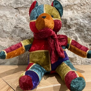 Könnte beinhalten: Ein Patchwork-Teddybär mit rotem Schal. Der Bär besteht aus Samtstoff in verschiedenen Farben, darunter Rot, Orange, Grün, Blau und Lila. Goldbesatz umrandet die Nähte. Der Bär sitzt auf einer Holzoberfläche.