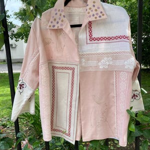 May include: A long-sleeved button-down shirt featuring a patchwork design of light pink fabric and white panels. The garment includes embroidered dandelion motifs, red geometric border stitching, and white lace trim details on the collar and cuffs.