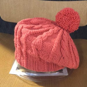 Puede incluir: Un gorro de punto de color coral con un diseño de punto trenzado y un gran pompón en la parte superior. El gorro está hecho de un hilo suave y texturizado. Un accesorio de invierno.