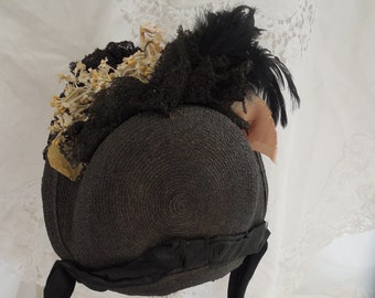 May include: A black straw hat with a black ribbon and black lace trim. The hat is decorated with a black feather and a cluster of white and yellow flowers.