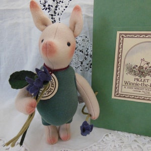 May include: A Piglet doll from Winnie-the-Pooh, holding purple flowers. The doll has a pink snout, ears, and cheeks, and wears a green outfit. A tag reads "GHT INC." and the box reads "PIGLET Winnie-the-Pooh R. JOHN WRIGHT DOLLS, INC."