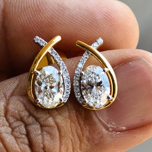 Puede incluir: Un par de pendientes de oro con piedras preciosas ovaladas y transparentes. Los pendientes tienen un marco dorado con un diseño entrecruzado y están adornados con pequeñas piedras transparentes.