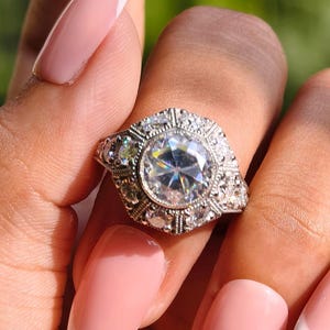 Puede incluir: Un anillo plateado con una piedra preciosa grande, redonda y transparente en el centro, rodeada de piedras más pequeñas. El anillo está en una mano con uñas francesas rosas y blancas. El fondo es verde borroso.