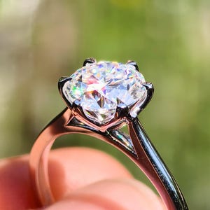 Könnte beinhalten: Nahaufnahme eines funkelnden Diamantrings. Der Ring hat einen großen, rund geschliffenen Diamanten, der in einem silbernen Band gefasst ist. Der Diamant reflektiert das Licht und erzeugt ein brillantes Funkeln. Der Ring wird vor einem verschwommenen grünen Hintergrund gehalten.