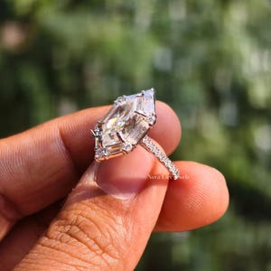 A breathtaking close-up of a NovaLux Jewels Dutch marquise-cut moissanite engagement ring held against a soft-focus nature background. The unique hexagon-modified marquise stone displays intense rainbow fire, flawlessly framed by a high-polish 14k white gold setting and a shimmering half-eternity pave band. This image highlights the "Architectural Luxury" aesthetic and high-end handcrafted quality of this modern bridal piece.