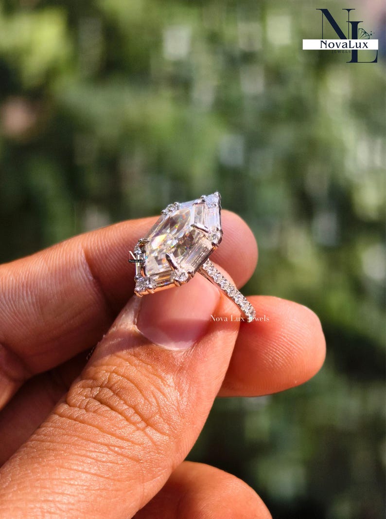 A breathtaking close-up of a NovaLux Jewels Dutch marquise-cut moissanite engagement ring held against a soft-focus nature background. The unique hexagon-modified marquise stone displays intense rainbow fire, flawlessly framed by a high-polish 14k white gold setting and a shimmering half-eternity pave band. This image highlights the "Architectural Luxury" aesthetic and high-end handcrafted quality of this modern bridal piece.