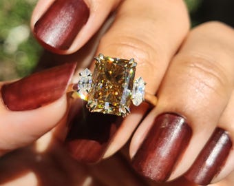 May include: A ring featuring a large, rectangular, radiant-cut brown gemstone set in the center, flanked by two smaller, marquise-cut clear gemstones on either side. The stones are mounted on a thin, polished gold-toned metal band.