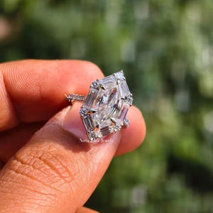 A romantic close-up of a sparkling moissanite engagement ring, symbolizing a modern love story that values both brilliance and ethical sourcing. The colorless D-color stone reflects only pure white light against the bright white gold setting. The soft-focus greenery in the background reinforces the brand's commitment to "Quiet Luxury" and sustainable, handcrafted elegance for the contemporary bride.