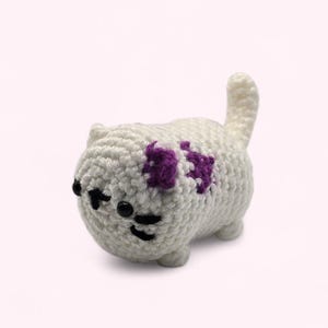 May include: A handmade, white crochet cat with a purple bow on its back. The cat has black eyes and whiskers. The cat is a small, round shape with a tail sticking up.