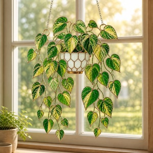May include: A stained glass hanging planter featuring a trailing plant design. The leaves are green and yellow, and the planter is white with a gold-coloured rim. The planter hangs from a chain in front of a window.