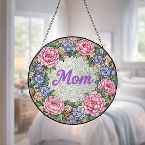 Puede incluir: Arte de ventana redondo de vidrieras con un diseño de corona floral. La corona presenta peonías rosas, flores azules y amarillas y hojas verdes. La palabra "Mom" está escrita en púrpura en el centro. La ventana cuelga de una cadena.