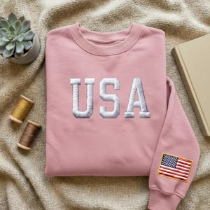 Könnte beinhalten: Ein hellrosa Sweatshirt mit dem Wort "USA" in weißen Blockbuchstaben gestickt. Der Ärmel hat eine kleine gestickte amerikanische Flagge. Das Sweatshirt wird auf einer neutralfarbenen Oberfläche präsentiert.