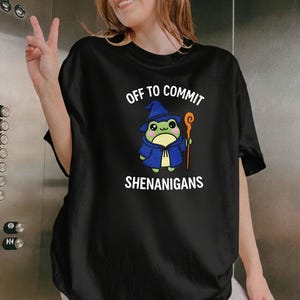 以下が含まれることがあります： 「OFF TO COMMIT SHENANIGANS」の文字と、魔法使いの衣装を着たカエルの漫画のイラストが描かれた黒いTシャツ。カエルは緑色、帽子とローブは青色、杖はオレンジ色です。テキストは白です。