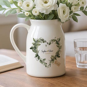 Peut inclure: Un pichet en céramique blanc avec une anse, rempli de fleurs blanches et de verdure. Le pichet présente un motif en forme de cœur avec du texte. Un verre d'eau et un livre ouvert sont sur une table en bois.