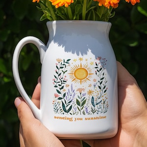 Puede incluir: Una jarra de cerámica blanca con asa, llena de flores de color naranja brillante. La jarra está decorada con un diseño de sol y flores, y las palabras "sending you sunshine". Las flores tienen centros de color marrón oscuro.