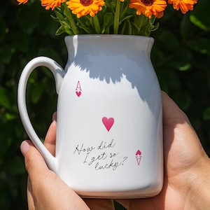 Puede incluir: Una jarra de cerámica blanca con asa, llena de flores naranjas vibrantes. La jarra tiene un diseño decorativo con símbolos de naipes y el texto "How did I get so lucky?"