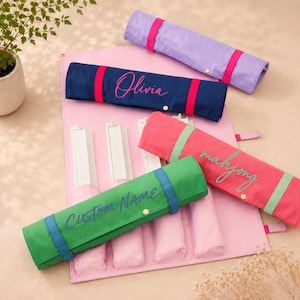 May include: A set of fabric mahjong tile racks and cases in lavender, navy blue, coral, and green. The navy case features the name Olivia in pink, the coral case says mahjong in light green, and the green case says CUSTOM NAME in blue.