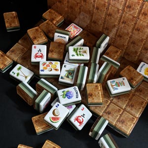 May include: A collection of Mahjong tiles featuring a mix of brown wood-grain backs and white faces. The faces display various illustrations including crabs, palm trees, tropical fruits, flowers, and the text JOKER, FLOWER, and letters E and N.