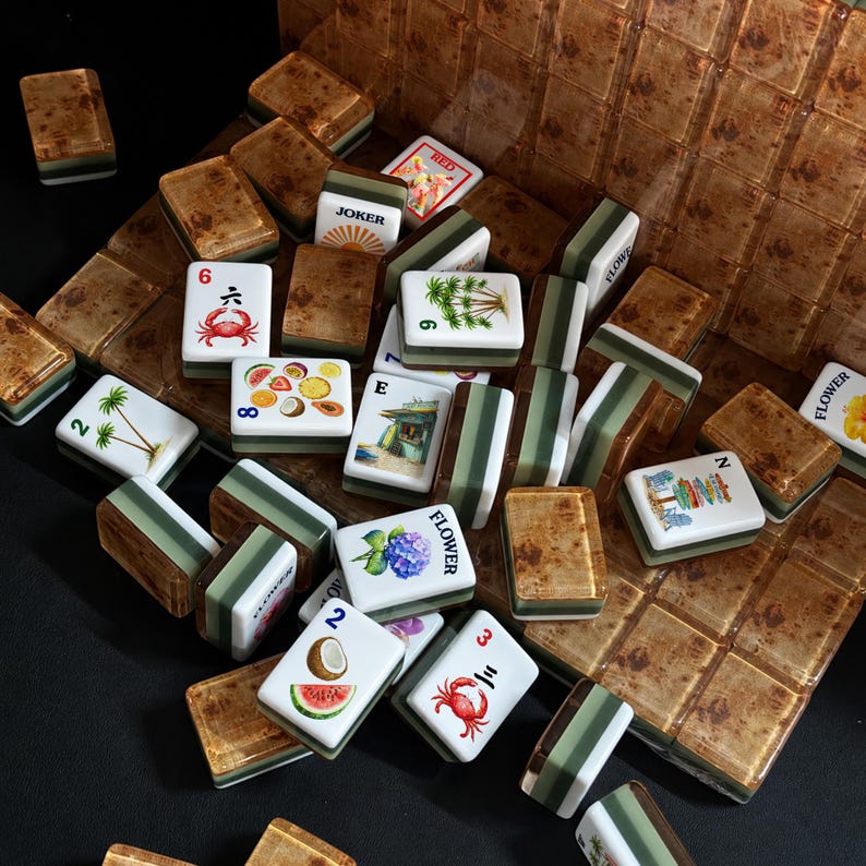 May include: A collection of Mahjong tiles featuring a mix of brown wood-grain backs and white faces. The faces display various illustrations including crabs, palm trees, tropical fruits, flowers, and the text JOKER, FLOWER, and letters E and N.