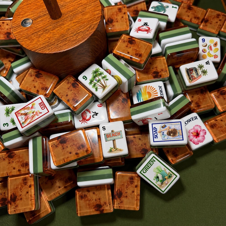May include: A collection of mahjong tiles featuring a faux wood grain pattern on the back and white faces with various colorful illustrations, including a beach sign, a hibiscus flower, a crab, palm trees, and text like RED, SOAP, and GREEN.
