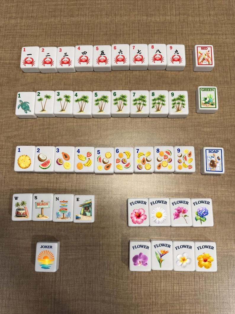 May include: A set of white rectangular mahjong tiles arranged in rows. The tiles feature colorful illustrations of crabs, palm trees, tropical fruits, flowers, beach scenes, and a joker tile. Each tile displays a number or text label.