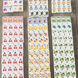 May include: A set of white rectangular mahjong tiles arranged in columns. The tiles feature colorful illustrations of crabs, tropical fruits, palm trees, sea turtles, flowers, beach scenes, and the word JOKER, along with numbers one through nine.