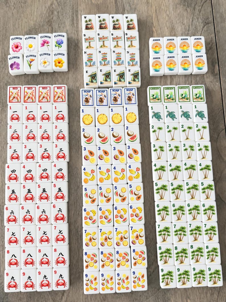 May include: A set of white rectangular mahjong tiles arranged in columns. The tiles feature colorful illustrations of crabs, tropical fruits, palm trees, sea turtles, flowers, beach scenes, and the word JOKER, along with numbers one through nine.