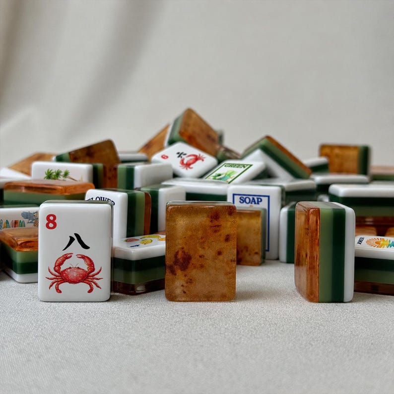 May include: A collection of rectangular mahjong tiles featuring a mix of white faces with red crab illustrations and the number 8, alongside translucent amber-colored resin backs with green and white layered bases. Some tiles display text like SOAP.
