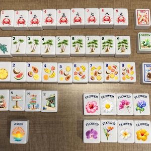 May include: A set of white rectangular mahjong tiles featuring colorful illustrations of crabs, palm trees, tropical fruits, beach scenes, and various flowers. Each tile displays a number or label, including Red, Green, Soap, W, S, N, E, Joker, and Flower.