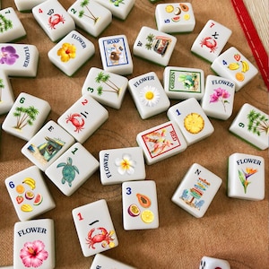 May include: A collection of white rectangular mahjong tiles featuring colorful illustrations of tropical flowers, fruits, crabs, palm trees, and beach scenes, along with two white dice with black and red pips scattered on a tan fabric surface.