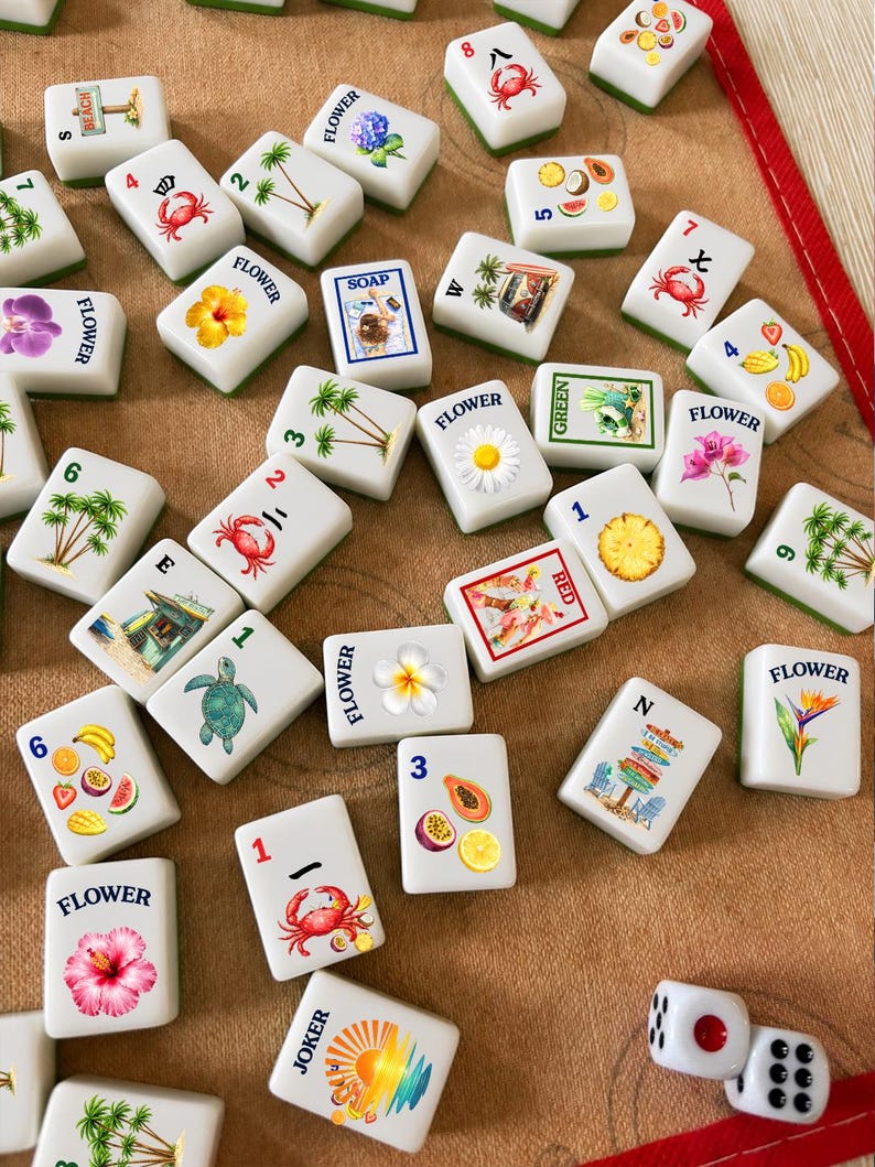 May include: A collection of white rectangular mahjong tiles featuring colorful illustrations of tropical flowers, fruits, crabs, palm trees, and beach scenes, along with two white dice with black and red pips scattered on a tan fabric surface.