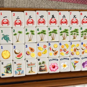 May include: A set of white mahjong tiles arranged in a box, featuring colorful illustrations of crabs, palm trees, tropical fruits, flowers, and beach scenes. Each tile displays a number or label, including Red, Green, Soap, Joker, E, N, S, W, and Flower.