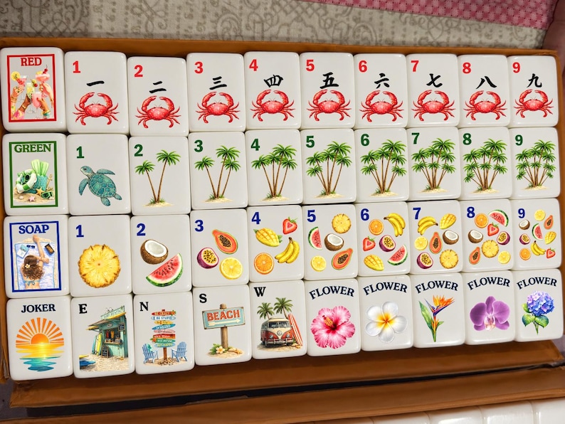 May include: A set of white mahjong tiles arranged in a box, featuring colorful illustrations of crabs, palm trees, tropical fruits, flowers, and beach scenes. Each tile displays a number or label, including Red, Green, Soap, Joker, E, N, S, W, and Flower.