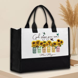 May include: Black and white tote bag with black handles. The bag features a design with sunflowers in mason jars, hummingbirds, and the text "God says you are..." and "Mrs. Megan."