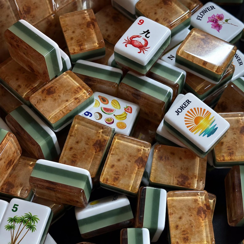 May include: A collection of mahjong tiles featuring a mix of designs, including a red crab with the number 9, a colorful fruit assortment, a sunburst graphic labeled JOKER, palm trees with the number 5, and a flower illustration with the text FLOWER.