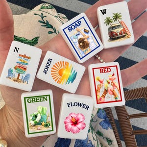 May include: A set of six white rectangular mahjong tiles featuring colorful illustrations and text. Designs include a beach scene with the word SOAP, a van with W, a signpost with N, a sun graphic with JOKER, a cocktail toast with RED, and a flower with FLOWER.