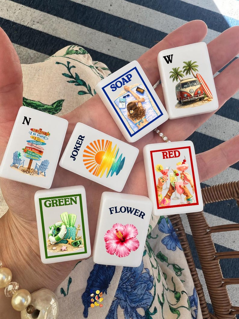 May include: A set of six white rectangular mahjong tiles featuring colorful illustrations and text. Designs include a beach scene with the word SOAP, a van with W, a signpost with N, a sun graphic with JOKER, a cocktail toast with RED, and a flower with FLOWER.